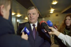 Mężczyzna w garniturze stoi przed ludźmi. Wokół niego są mikrofony. Po prawej stronie stoi lonbieta a onok widać rękę bez twarzy. Po lewej stronie jest mężczyzna, który stoi tyłem do obiektywu a jego sylwetka nie jest w pełni pokazana.