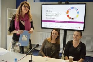 Za stołem w sali siedzą dwie kobiety po prawej stronie a jedna stoi po lewej stronie i trzyma teczkę. Przed sobą mają mikrofon, kubek plastikowy. Za nimi wyświetlana jest prezentacja pt.” 12 edycja Światowego Tygodnia Przedsiębiorczości”.