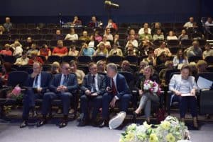 Osoby siedzą na widowni. W pierwszym rzędzie siedzą mężczyźni ubrani w garnitury.