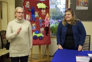 Dwie kobiety stoją w sali. Za nimi na tablicy przyczepione są kukiełki materiałowe. Na stole po prawej stronie leżą broszury.