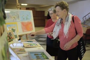 Dwie starsze kobiety stoją przed stołem na którym są obrazy. Jedna z nich ma wyciągniętą rękę i palec skazujący. Kobieta za nią patrzy na stół. Obok niej na sztaludze stoi rama z czterema obrazami. Obie panie stoją bokiem