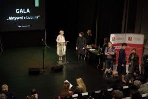 Na scenie przy mównicy stoi kobieta. Jest zwrócona w prawą stronę gdzie stoi kobieta. Po prawej stronie stoi grupa osób a w pierwszym rzędzie siedzą osoby tyłem do obiektywu.