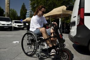 Mężczyzna siedzi na wózku inwalidzkim. Jest na deptaku w mieście. Obok niego jest fragment busa.