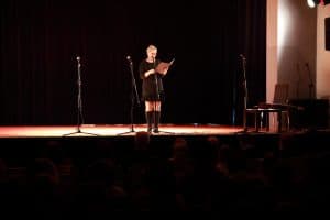 Kobieta stoi na scenie. W rękach trzyma otwartą teczkę, a mikrofon ma blisko twarzy. Obok niej stoją dwa statywy.