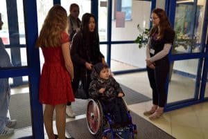 Kobieta wjedżdza przez drzwmi otwierane automatycznie z dzieckiem na wózku inwalidzkim. Po bokach przy drzwaich stoja kobiety.