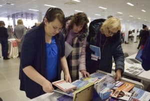 Ludzie pochylają się nad stołem z broszurami i ulotkami. W tle inni ludzie na sali.