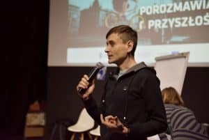 Mężczyzna w średnim wieku trzyma mikrofon przy ustach. Za nim wyświetlana jest prezentacja „Porozmawiajmy o przyszłości miasta”. Na fotografii znajduje się także kobieta, która jest uchwycona tyłem do kamery.