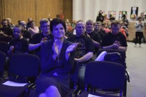 Ludzie siedzą na krzesłach w rzędach. Kobieta uśmiecha się i trzyma ręce w geście klaskania.