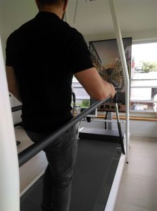 Mężczyzna stoi na bieżni, ręce trzyma na poręczach. Na ekranie wyświetla się obraz z budynkami i drogą.