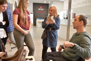 Kobiety stoją w pomieszczeniu. Jedna z nich trzyma telefon i patrzy na drugą. Po prawej stronie siedzi mężczyzna na wózku inwalidzkim.