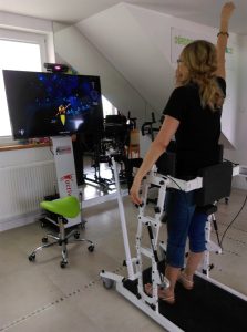 Kobieta stoi na urządzeniu rehabilitacyjnym i patrzy w ekran. Na ekranie przed nią wyświetla się obraz.