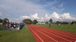 Grupa osób jest na boisku. Po lewej stronie siedzi widownia. Między widownią a boiskiem jest bieżnia.