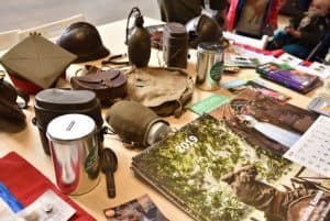 Na stole leżą puszki, kalendarze wojskowe, granaty, czapki, manierki.