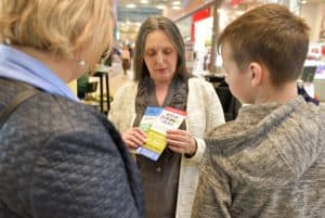 Kobieta trzyma w rękach dwie ulotki. Po prawej i lewej stronie stoi chłopiec i kobieta. Są oni tyłem do obiektywu.