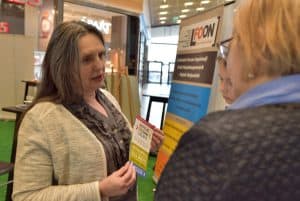 Kobieta stoi z ulotkami bokiem do chłopca i kobiety. Kobieta stoi tyłem do obiektywu.