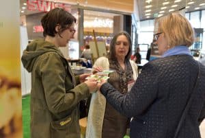 Trzy kobiety stoją naprzeciwko siebie. Dwie z nich trzymają ulotkę, a trzecia podaje następną.