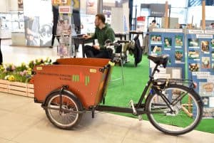 Na płytkach stoi rower. Z przodu ma zamontowaną dużą skrzynkę. Za nim stoją tablice ze zdjęciami oraz siedzi mężczyzna.