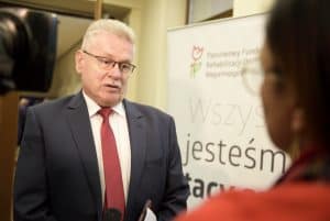 Mężczyzna ma twarz zwróconą w kierunku kobiety w tle. W tle także baner reklamowy “Państwowy Fundusz Rehabilitacji Osób Niepełnosprawnych”