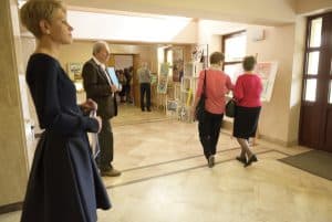 W pomieszczeniu jest kilka osób. Dwie panie idą i są tyłem do obiektywu. Po lewej stronie stoi kobieta i trzyma ręce lekko ugięte przy ciele. Za nią stoi mężczyzna.