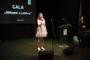Na scenie jest młoda dziewczyna, która trzyma mikrofon w ręce. Po prawej stronie stoi tłumaczka migowa. Z tyłu napis: “ Gala Aktywni z Lublina”.