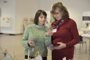 Na zdjęciu są dwie kobiety, które stoją. Jedna trzyma w dłoni kwiatek z papieru.