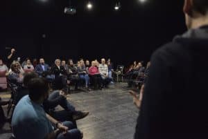 Ludzie siedzą w dużym kole na krzesłach. Po prawej stronie stoi mężczyzna, który ukazany jest od tyłu. Środek dłoni kieruje ku górze.