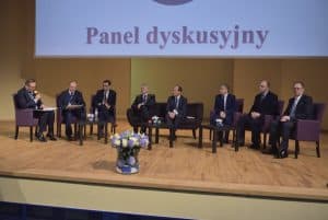 Grupa osób siedzi na scenie w fotelach. Mężczyzna po lewej stronie trzyma mikrofon i ma teczkę na kolanach. Nad nimi wyświetla się napis “ Panel dyskusyjny.”