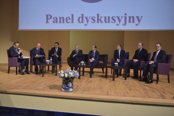 Grupa osób siedzi na scenie w fotelach. Mężczyzna po lewej stronie trzyma mikrofon i ma teczkę na kolanach. Nad nimi wyświetla się napis “ Panel dyskusyjny.”