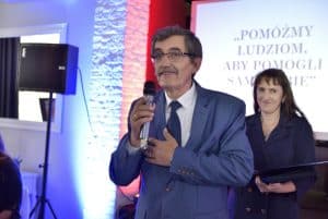 Mężczyzna stoi na scenie. W dłoni ma mikrofon. W tle za nim kobieta, która się uśmiecha.