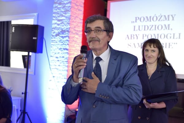 Mężczyzna stoi na scenie. W dłoni ma mikrofon. W tle za nim kobieta, która się uśmiecha.