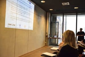 Osoby siedzą w sali a obok nich są położone kartki. Na ścianie widnieje prezentacja z tektem “ ułatwienia architektoniczne-42%; dostosowanie toalety- 39%; zapewniony swobodny dostęp do stanowiska pracy (brak barier tj. nierówności podłogi)-37%; brak potrzeby dostosowanie stanowiska pracy- 27%; możliwość dostosowania wysokości stołu/ biurka do potrzeb osoby niepełnosprawnej- 24%.