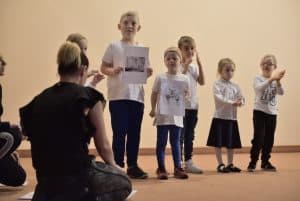 Na scenie stoją dzieci. Po lewej stronie jest kobieta, która siedzi twarzą do dzieci,a tyłem do obiektywu. Dwoje dzieci trzymają przed sobą kartki z rysunkami.