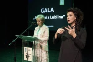 Na scenie są dwie kobiety. Po lewej stronie stoi za pulpitem kobieta, która ma blisko ust mikrofon. Po prawej stronie jest tłumaczka języka migowego. W tle prezentacja z napisem: “Gala”.