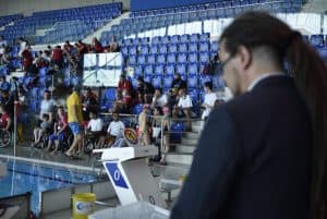 Po prawej stronie stoi mężczyzna w garniturze. Jest on tyłem do obiektywu. przed nim siedzą ludzie na trybunach.