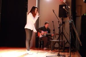 Na scenie stoi kobieta z mikrofonem. Za nią siedzi mężczyzna z gitarą. Jest ona bokiem do obiektywu.