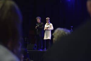 Dwie osoby stoją na scenie- kobieta i mężczyzna. Oboje trzymają mikrofony. Ludzie siedzą na widowni i są tyłem do obiektywu