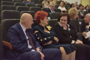 Na widowni siedzi grupa osób. Kobieta po prawej stronie jest odwrócona w stronę innej kobiety i mężczyzny.