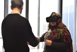 Mężczyzna i kobieta stoją naprzeciwko siebie. Kobieta ma na sobie duże Gogle VR. Mężczyzna trzyma kobietę za rękę.