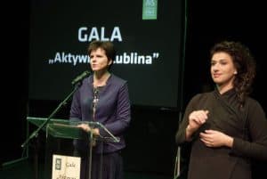 Kobieta po lewej stronie stoi za pulpitem i ma mikrofon blisko ust. Po prawej stronie jest tłumaczka języka migowego. Na prezentacji za nimi jest napis: “ Gala. Aktywni z Lublina”.