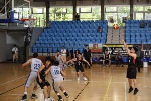 Po prawej stronie stoi mężczyzna z wyciągniętymi rękami do góry. Nad nim jest piłka w powietrzu. Pod koszem grupa mężczyzn.