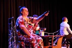 Na widowni siedzi grupa osób. Kobieta ma wyciągniętą wysoko rękę i trzyma w niej pilota od telewizora. Obok niej siedzą mężczyźni.