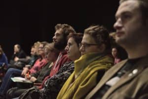 Publiczność jest sfotografowana bokiem. Na widowni siedzą: mężczyzna, dwie kobiety i kolejny mężczyzna. Wszyscy są ustawieni bokiem do obiektywu.