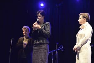 Trzy kobiety stoją na scenie. Jedna z nich trzyma mikrofon blisko ust.