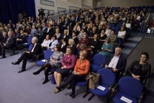 Na widowni siedzi duża grupa osób. Niektóre miejsca siedzące są puste i jest na nich położona kartka. Wszyscy są bokiem do obiektywu.