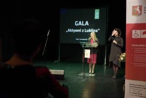 Dwie kobiety stoją na scenie. Po lewej stronie kobieta stoi przy pulpicie i ma mikrofon blisko ust. Po prawej jest tłumaczka języka migowego. Na prezentacji za nimi jest napis: “ Gala. Aktywni z Lublina”.
