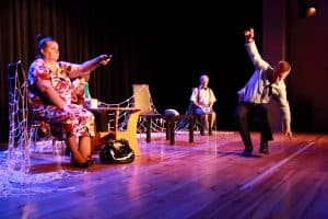 Na scenie siedzi grupa osób. Kobieta po lewej stronie ma wyciągniętą rękę. Na scenie są także krzesła i stoliki. Sieć rybacka jest rozciągnięta na krzesła.