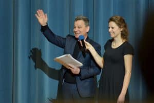 Mężczyzna i kobieta stoja na scenie. Kobieta trzyma mikforon który wyciąga w w stronę męzyczyzny. Męzczyzna podnosi ręke ku górze.