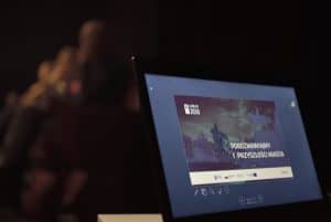 W prawym dolnym rogu widnieje laptop. Na monitorze wyświetlana jest prezentacja pt. „ Porozmawiajmy o przyszłości miasta”. W tle widać zaledwie zarys siedzących ludzi.