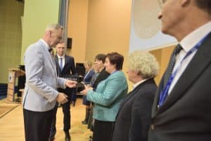 Na scenie stoi grupa osób. Mężczyzna stoi naprzeciwko kobiety i trzyma przed nią statuetkę. Kobieta ma wyciągnięte ręce w jej kierunku.