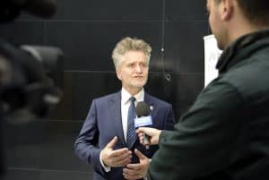 Starszy mężczyzna w garniturze mówi do mikrofonu, który trzyma inny mężczyzna. dziennikarz stoi bokiem do obiektywu. Po lewej stronie jest część kamey.